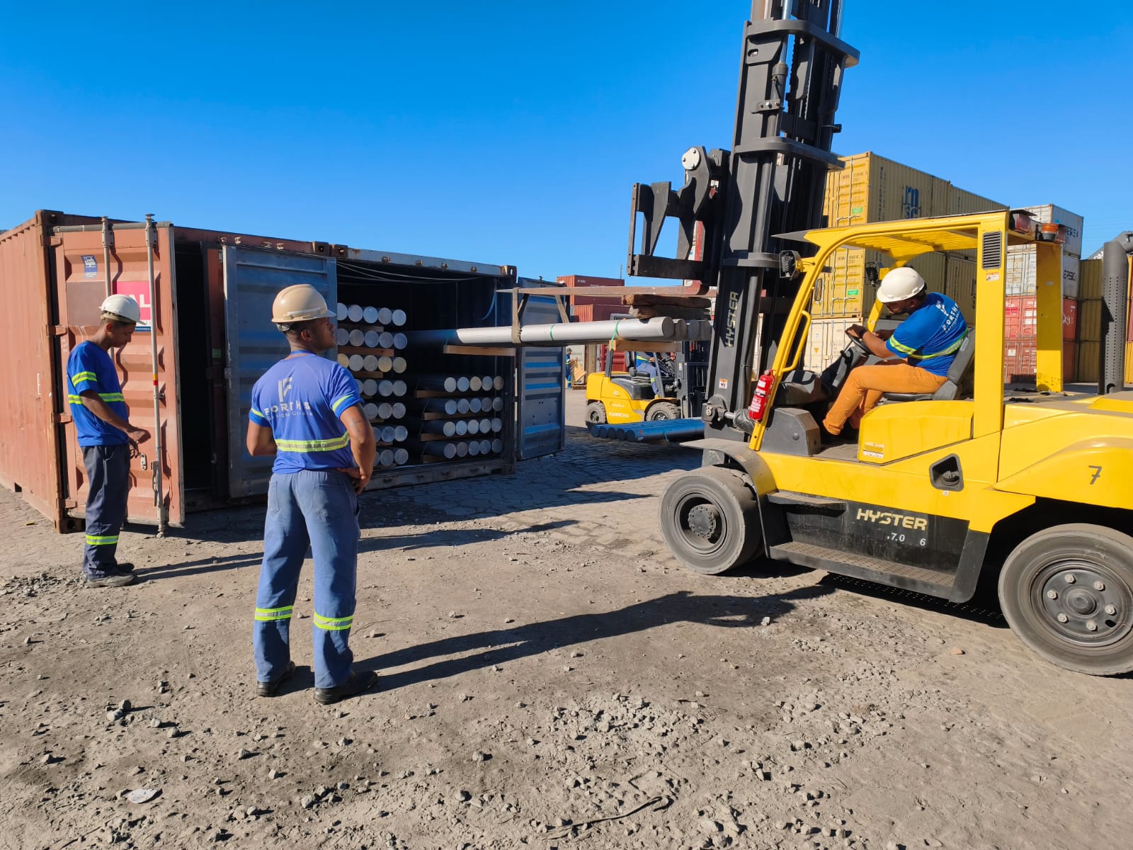Operador de empilhadeira Hyster movimentando tubos ao lado de contêiner com equipe Forths acompanhando